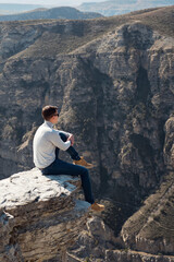 Guy in denim jeans and white shirt sits on edge of steep rocky cliff against large brown bare mountains under sunlight on autumn day.