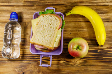 Lunch box with sandwiches, bottle of water, banana and apple on a wooden table. Top view