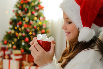 Young girl wearing white fluffy sweater holding a cup of cacao with bunch of marshmallows at home. Christmas tree with stack of presents underneath, festive bokeh light. Blurry background, copy space