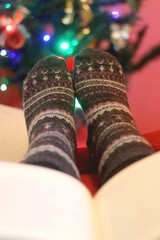 Feet in warm socks and open book by the Christmas tree. Selective focus.