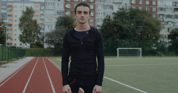 Portrait of disabled guy with prosthetic running blades standing at sports field. Young amputee male runner looking to camera outdoors. Concept of motivational sports footage.