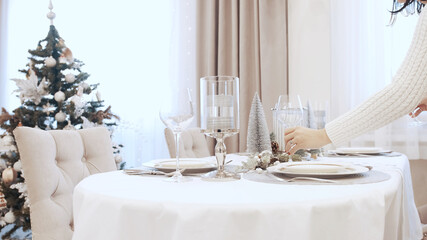 A girl in a white dress serves wine glasses at the Christmas table. Festive table setting