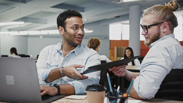 Indian male working at notebook on business project, sitting at table in office, caucasian colleague giving him folder with documents