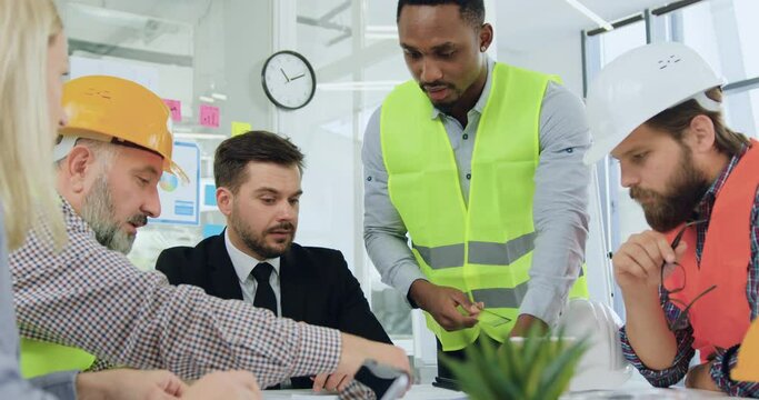 Good-looking confident professional black-skinned engineer explaining building details on blueprint to his serious high-skilled bearded manager during meeting with anothers members of working team