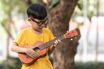 An Asian little boy wearing glasses is happily playing the ukulele. Asian little child is trying to play the ukulele with a fully happy moment.