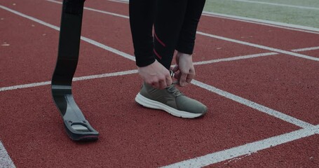 Crop view of disabled male person with prosthetic running blades tying shoelaces and standing at sports field. Concept of motivational sports footage, handicap,paralympian. - Powered by Adobe