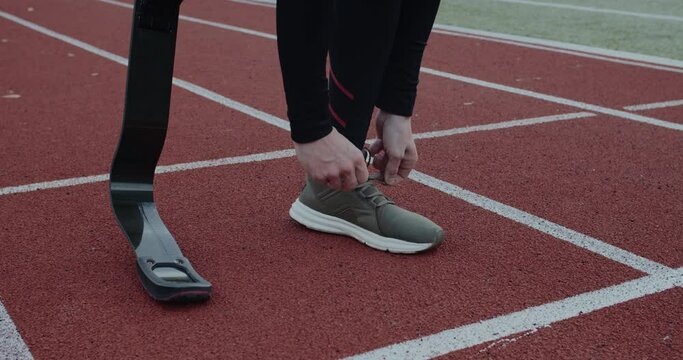 Crop view of disabled male person with prosthetic running blades tying shoelaces and standing at sports field. Concept of motivational sports footage, handicap,paralympian.