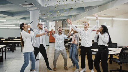 Group of diverse colleagues in party hats are blowing in whistles, smiling and dancing celebrating birthday party