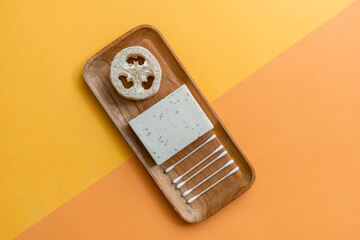 Top view luffa sponge, solid shampoo and bamboo ear sticks on wooden tray on yellow and orange...