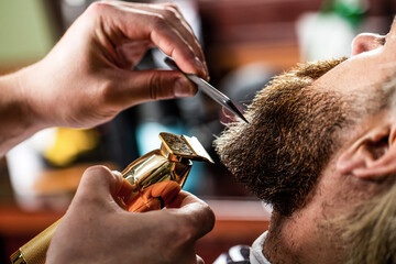 Barber works with a beard clipper. Hipster client getting haircut. Hands of a hairdresser with a beard clipper, closeup - obrazy, fototapety, plakaty