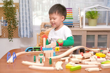 Cute little Asian boy child having fun playing with wooden train, railway and building block toys indoor at home, photo in real life interior