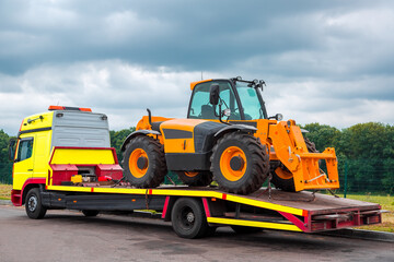 The tow truck on the platform transports the new tractor, equipment for technical and agricultural works stands on an asphalt road near the curb, in the background a forest in cloudy weather, nobody.