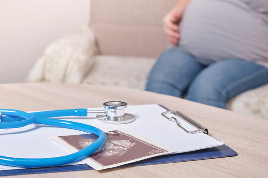 stethoscope and ultrasound scan on the table, pregnant woman sitting on the sofa on the background