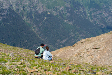 Fototapeta premium Rear view of man and woman sitting on rock and resting in mountains. Married couple enjoy beautiful view of mighty mountains in sunny weather.