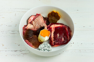 Natural healthy dog food in bowl on white wooden background. BARF Diet. Raw beef meat, cheeks, egg cottage cheese and food supplements.
