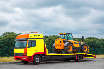 The tow truck on the platform transports the new tractor, equipment for technical and agricultural works stands on an asphalt road near the curb, in the background a forest in cloudy weather, nobody.