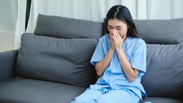 Sick Asian woman with flu, cold, fever and cough sitting on couch at home. Ill person blowing nose and sneezing with tissue and handkerchief. medicine. Infection in winter. Resting on sofa.