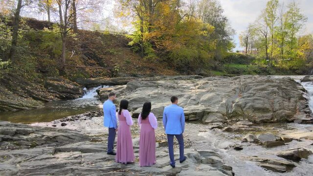 Group of four people, two men in suits and two women and dresses standing near small mountain river in autumn warm day.