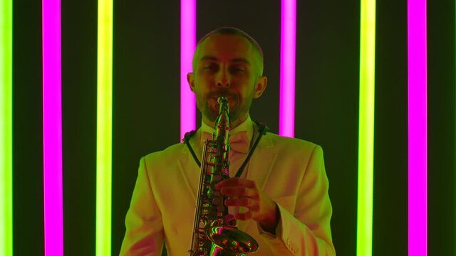 A man in a suit is masterly performing a composition on the saxophone. The musician plays a musical instrument in the studio against the background of multicolored neon tubes. Close up, slow motion.