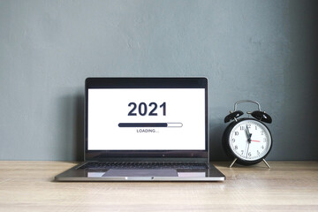 Loading the new year 2021 on a laptop screen with alarm clock countdown on a wooden table with grey background. 