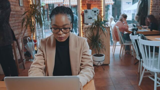 Smart concentraed young afro-american woman wearing eyeglasses looking on computer screen working with laptop inside cafeteria.