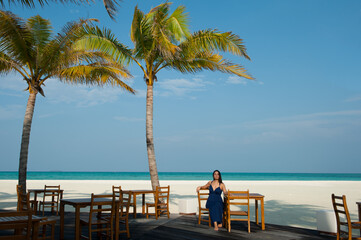 Beautiful brunette woman in blue dress sitting on the shore restaurant . White sand and turquoise ocean view