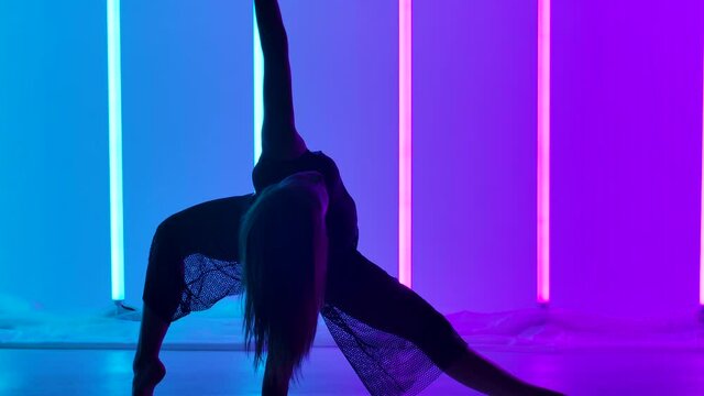 A silhouette of a girl dancer performing acrobatic elements of contemporary choreography. A artist is dancing in a studio against a background of blue and pink neon tubes in slow motion. Close up.