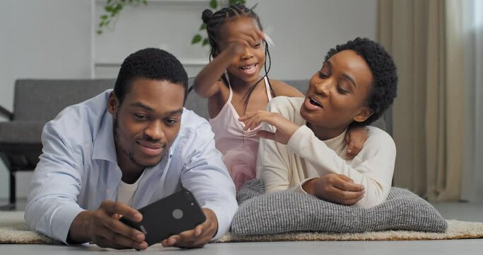 Afro american ethnic family taking photo on mobile phone camera posing having fun lying together looking at screen of smart phone three people spending time at home interior, love and support concept