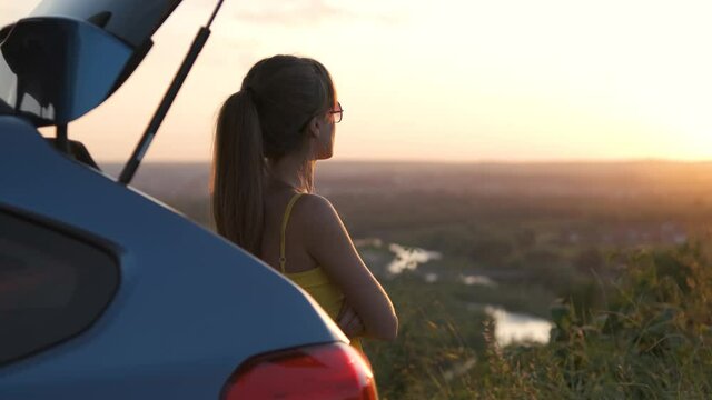 Young woman in summer dress standing near her car enjoying sunset view. Travel and vacations concept.