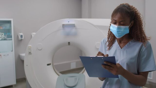 Portrait of black female tech in face mask writing on clipboard in CT scan room in hospital, then looking at camera