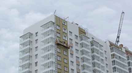 Fototapeta premium Construction of a multi-storey building. Suspended cradle with a worker. Wall insulation and mounting of a ventilated facade.