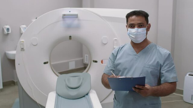 Portrait Of Arab Male Tech In Face Mask Standing In CT Scan Room In Hospital And Writing On Clipboard, Then Looking At Camera