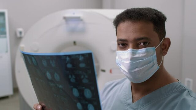PAN shot of Arab male doctor in face mask standing in radiology room and studying MRI scan of patient, then looking at camera