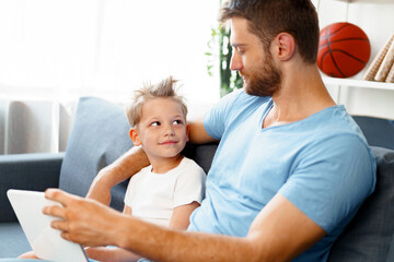 Little boy and his dad watching videos on digital tablet together sitting on sofa
