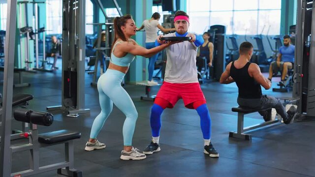 Caucasian athletic strong woman helping weak beginner man sportsman lifting barbell discs training lesson together in professional gym club. People train on background.