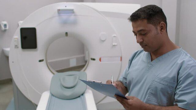 PAN shot of Arab male tech standing in room with CT scanner and writing on clipboard