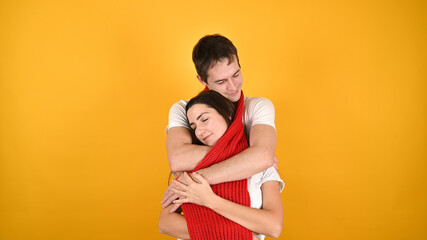 Young loving couple wrapped themselves in a long warm red scarf. Young man and woman hugs on yellow background with copy space. Valentine's day concept.