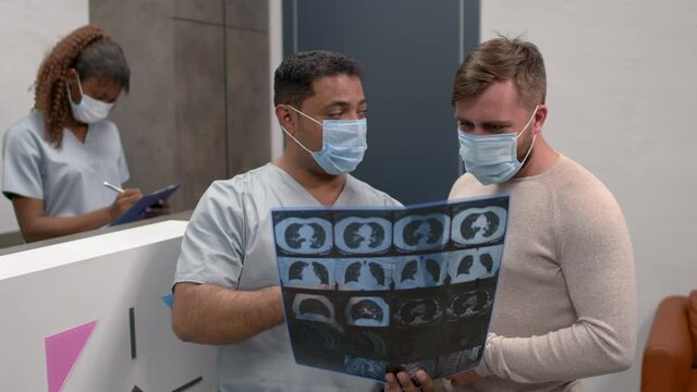 Medium shot of male doctor and patient in face masks looking together at chest CT scan in reception area of hospital