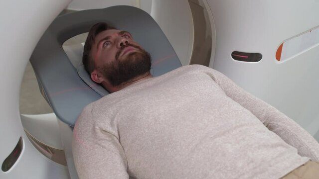 Medium shot of bearded male patient lying still on treatment couch inside CT scan