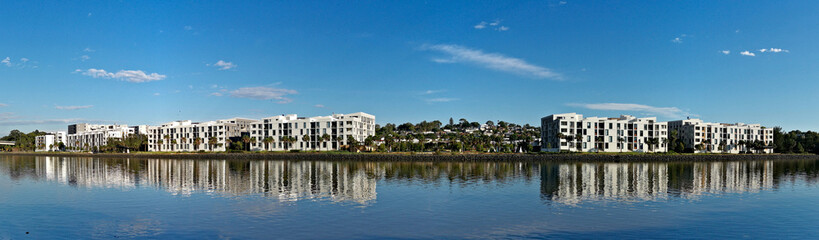 Fototapeta na wymiar Beautiful panoramic view of a river with reflections of modern apartment buildings, blue sky and trees on water, Parramatta river, Wilson Park, Silverwater, Sydney, New South Wales, Australia 