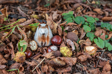 Small porcelain hedgehog on the ground with fallen autumn leaves and chestnut