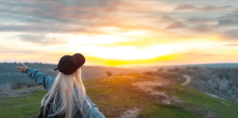 Fototapeta na wymiar Back view of young happy blonde girl with black cap, on peak of hills at sunset. Travel concept. Panoramic photo with copy space.
