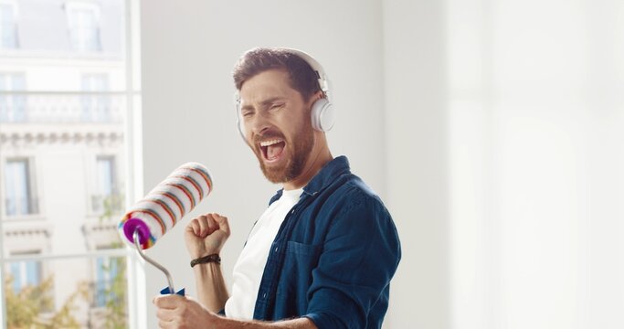 Close up of cheerful Caucasian funny man listening song in headphones, dancing, moving and singing to roller brush during home repair. Renovation and improvement Male having fun while renovating house