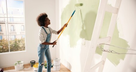 Young African American woman dancing and painting wall with roller brush while renovating...