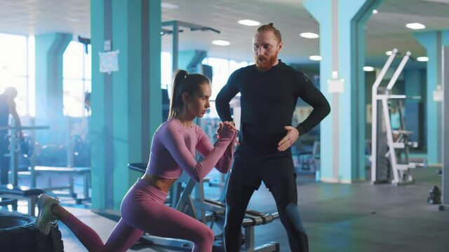 In gym female bodybuilder attractive woman talking with personal trainer professional male doing intense training squats exercise working out together. Sports.