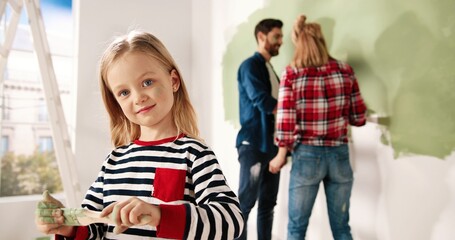 Close up portrait of adorable happy little cute girl smiling to camera standing in room with paint brush in hands. Mom and dad painting wall in green colour on background. Home repair. Interior design