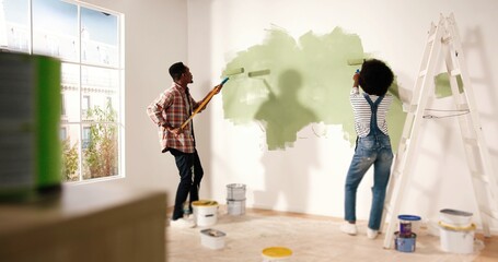 Back view of young lovely African American family married couple doing renovating works in house. Wife and husband painting walls together using roller brushes redecorating and redesigning home - Powered by Adobe