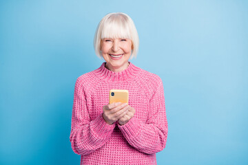 Photo portrait of smiling old woman holding phone in two hands isolated on pastel blue colored background