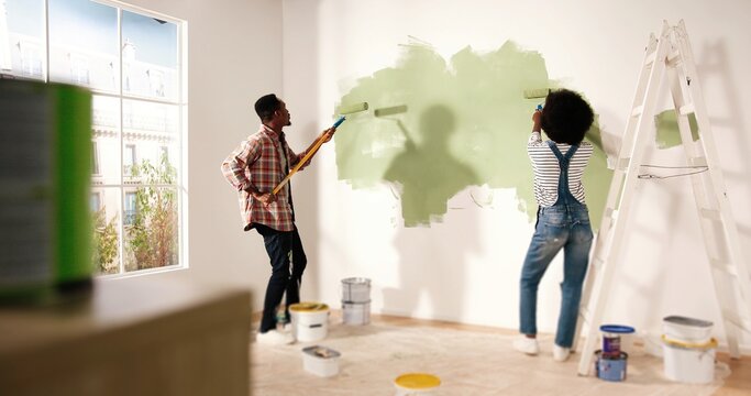 Back view of young lovely African American family married couple doing renovating works in house. Wife and husband painting walls together using roller brushes redecorating and redesigning home