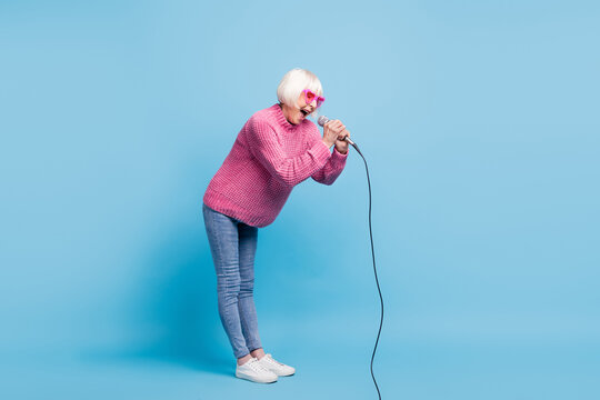 Full length photo portrait of screaming old lady holding microphone in two hands isolated on pastel blue colored background